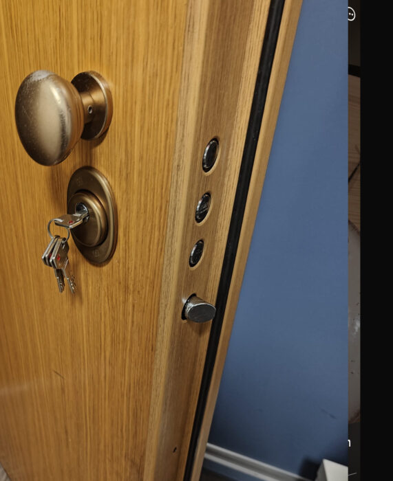 Puerta acorazada de madera clara abierta mostrando pomo, bombín con llaves de punto rojo y cuatro puntos de anclaje vistos desde el interior
